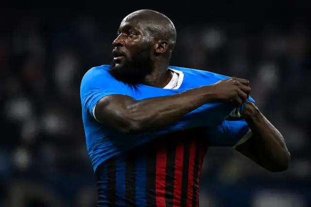 Lukaku si toglie la maglia del Napoli e sotto si intravede metà maglia Inter e metà maglia Milan, immagine creata con intelligenza artificiale